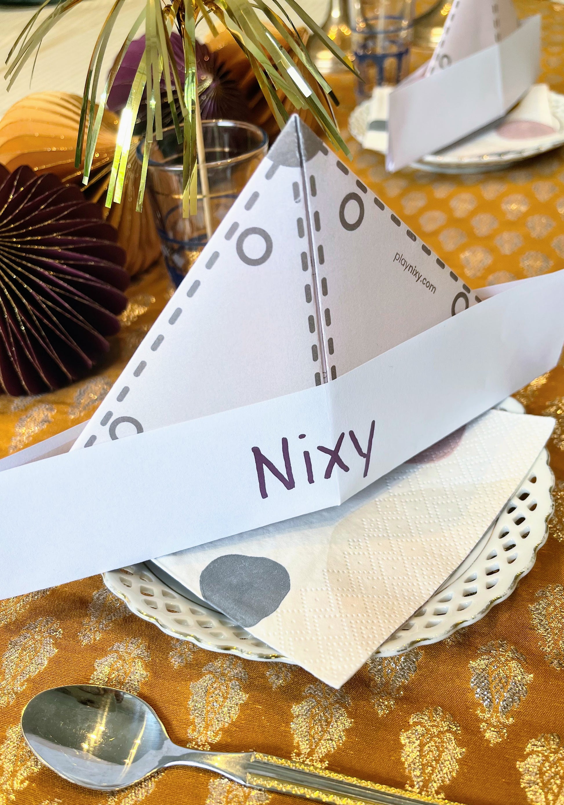 Safari hat place card with 'Nixy' on a decorative table setting with gold tablecloth and silver spoon.